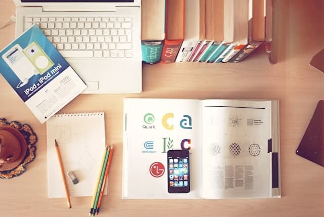 Top-down view of a study desk with a laptop, business textbooks, phone, notebook with pencils, and marketing brochures representing a learning environment for Business English.