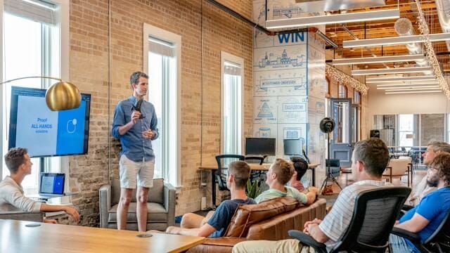 A man delivering a business learning session in a modern office while a group of professionals listen attentively. Using a visual aid to present in a collaborative and informal business training environment.