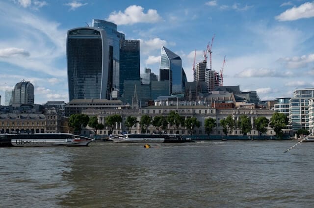 View of London’s financial district skyline from across the River Thames, featuring iconic modern skyscrapers under a blue sky.