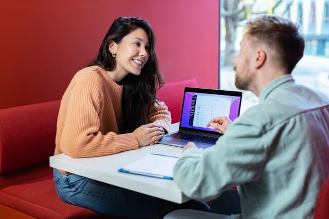 Two professionals having a business conversation at a modern office booth. One is taking notes on a clipboard while the other works on a laptop displaying a platform and practicing Business English communication skills.