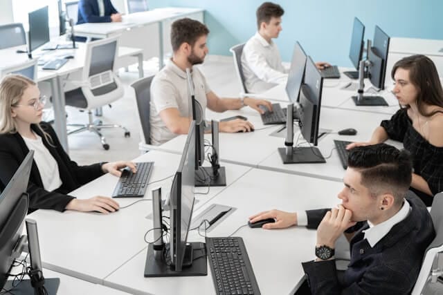 A group of young professionals sits at individual desks in a bright, modern computer lab, each focused on their desktop monitors. The atmosphere reflects a structured learning environment, ideal for education and e-learning.
