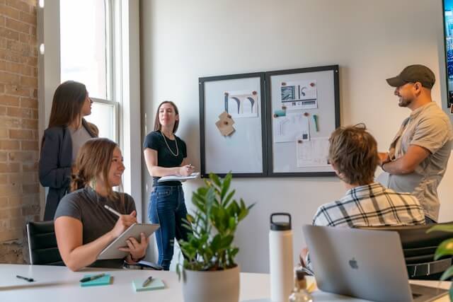 Group of professionals engaged in a training session a modern office. One woman stands and presents information pinned on a board while learners at a table take notes and interact.