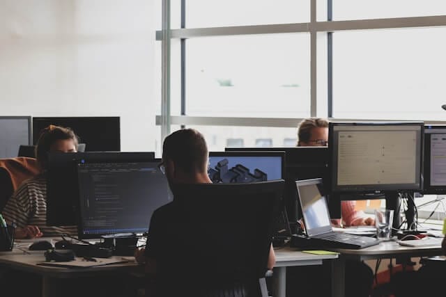 Office environment with multiple professionals working at computer stations in a modern, collaborative workspace with natural light, showing individuals engaged in work tasks.