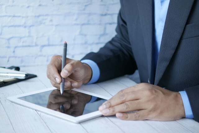 Business professional in a dark suit using a stylus to write on a digital tablet at a white desk. The image captures effective management communication in professional environments.